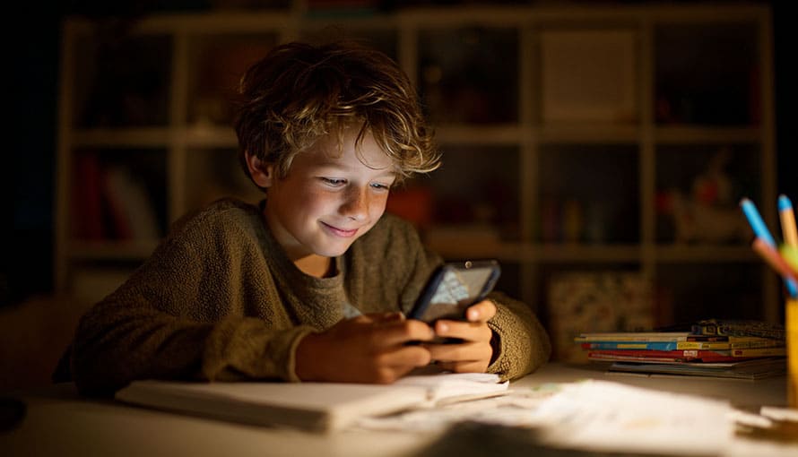 Chlapec sedí v prítmí svojej izby za stolom a pozerá do mobilu ktorý mu ožaruje tvár